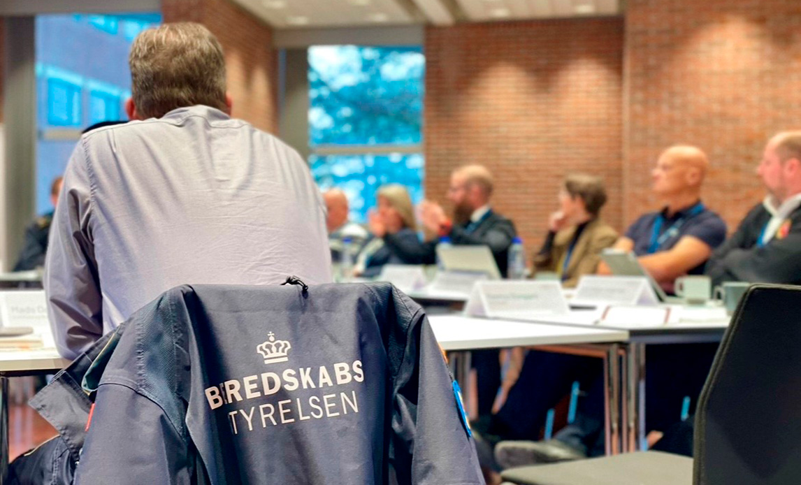 Billedet viser i baggrunden deltagerne der diskuterer emner under workshoppen. I forgrunden sidder en deltager fra Beredskabsstyrelsen, hvilket kan ses da hans jakke, som er i fokus, læser 'Beredskabsstyrelsen'.